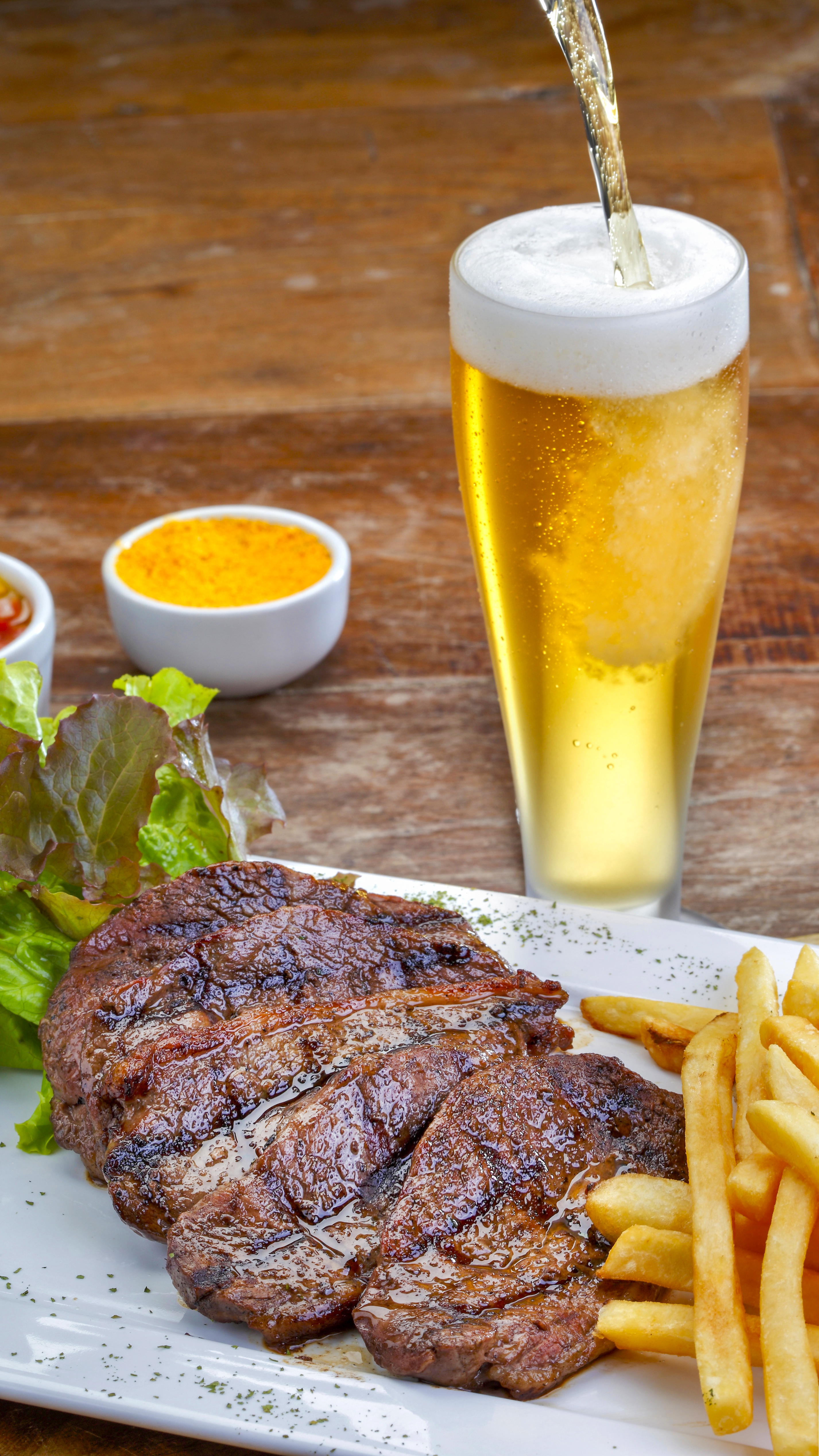 Delicious grilled picanha steak with french fries, salad, farofa, and a cold pouring beer on a rustic wooden table. Traditional Brazilian bbq.