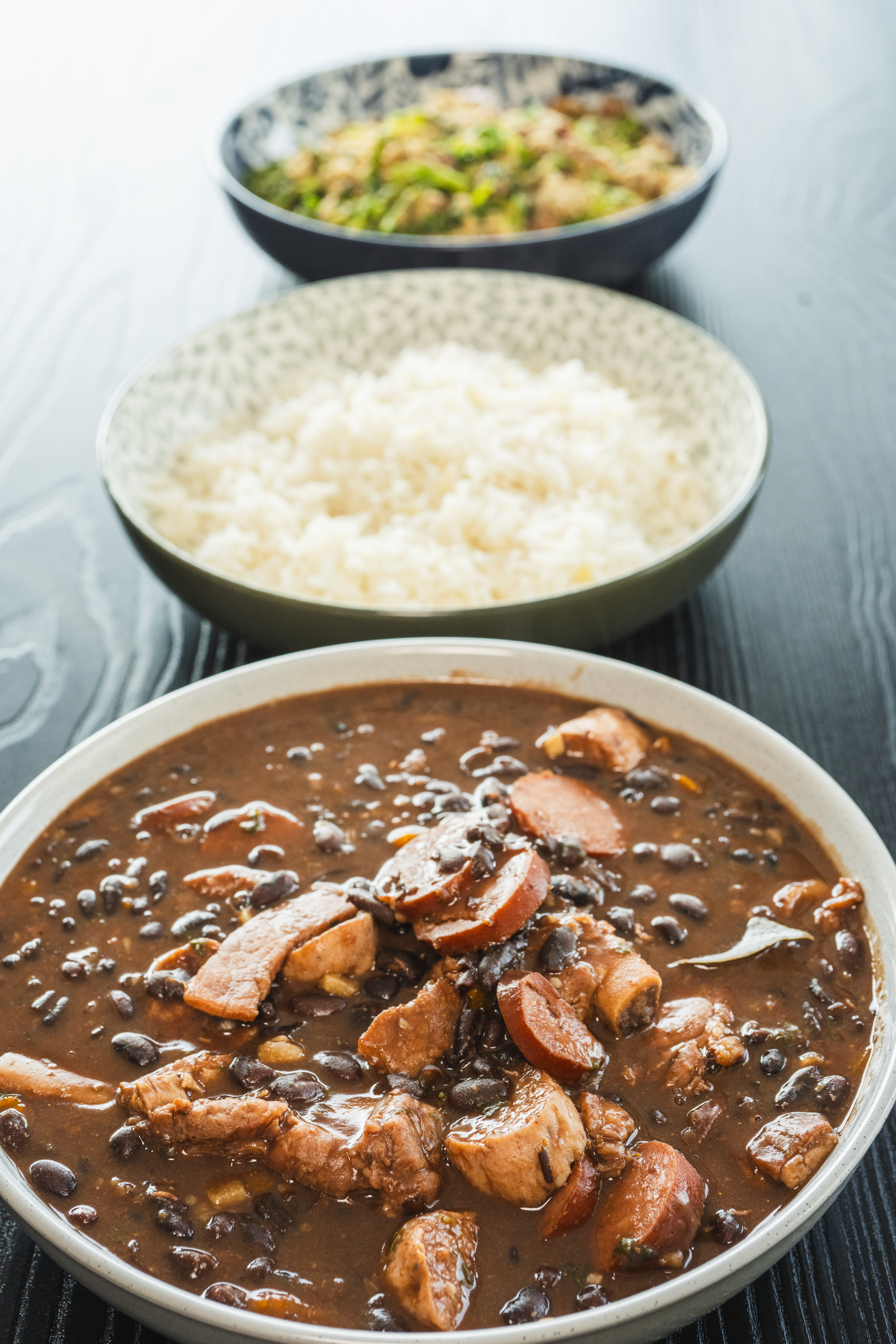Homemade Brazilian feijoada made with black beans and pieces of pork, served in bowls with white rice and toasted cassava flour. Traditional Brazilian lunch prepared at home.
